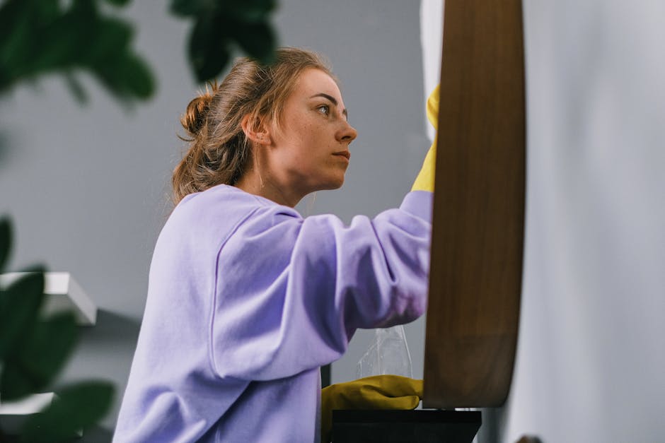 A young woman with light brown hair tied back, wearing a lilac long-sleeve shirt and yellow cleaning gloves, is attentively deep cleaning a wooden surface, possibly a table or cabinet, in a well-lit i