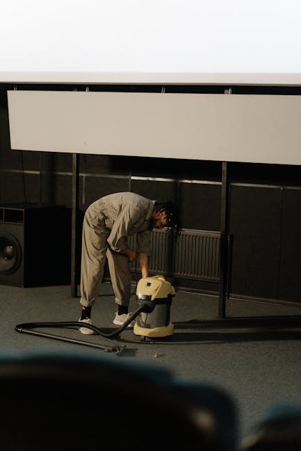 A person is kneeling on a patterned carpet in a room with warm lighting, using both hands to load a white cloth into a yellow and black vacuum cleaner. The floor is a traditional patterned rug, and th